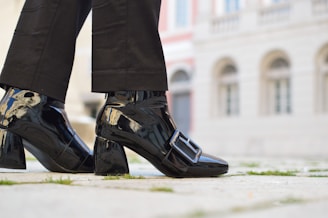 A person is wearing shiny black patent leather shoes with a large buckle detail. The shoes have a block heel and are paired with black pants. The background features the facade of a building with stonework and several arched windows, creating an urban setting.