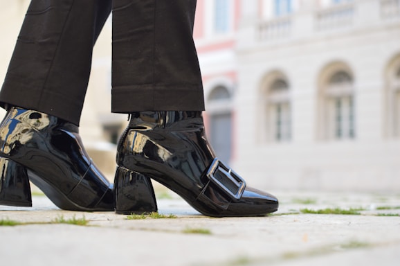 A person is wearing shiny black patent leather shoes with a large buckle detail. The shoes have a block heel and are paired with black pants. The background features the facade of a building with stonework and several arched windows, creating an urban setting.