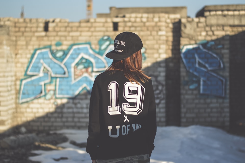 woman wearing black and white jacket and black fitted cap facing gray concrete brick wall with blue graffiti at daytime