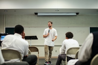 A man in a white t-shirt and khaki pants is speaking into a microphone in a classroom setting. He stands in front of a whiteboard with various words written on it. Several seated individuals are listening attentively, dressed in formal attire.