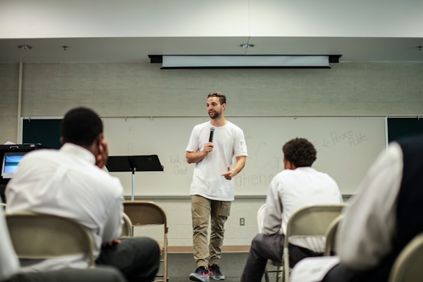 A man in a white t-shirt and khaki pants is speaking into a microphone in a classroom setting. He stands in front of a whiteboard with various words written on it. Several seated individuals are listening attentively, dressed in formal attire.