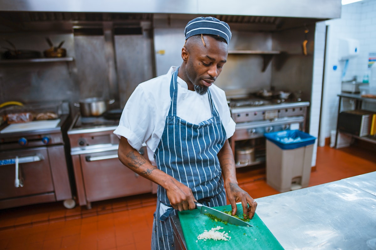 A chef in a striped apron and cap chopping herbs on a green board in a stainless-steel professional kitchen