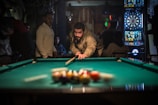 A person is lining up a shot on a pool table in a dimly lit room, surrounded by others who are standing or sitting. Dartboards are visible in the background, adding to the recreational setting.
