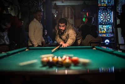 A person is lining up a shot on a pool table in a dimly lit room, surrounded by others who are standing or sitting. Dartboards are visible in the background, adding to the recreational setting.
