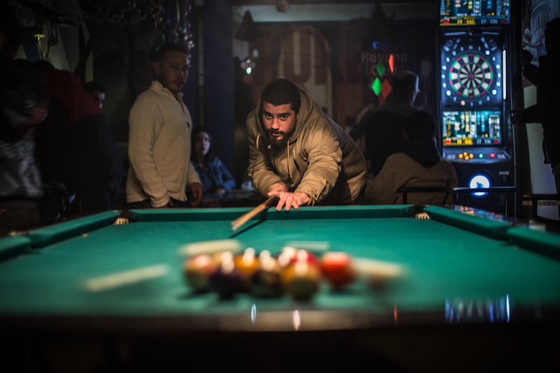 A person is lining up a shot on a pool table in a dimly lit room, surrounded by others who are standing or sitting. Dartboards are visible in the background, adding to the recreational setting.