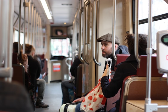 Inside a public transportation vehicle with passengers seated. A man wearing a flat cap looks attentively while sitting beside a person holding a white bag with red dots. The interior is lined with vertical metal poles and has a row of red seats.