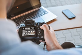 A person is holding a DSLR camera with a close-up view of the camera's screen, which displays various settings. In the background, a laptop is partially visible on a desk. A smartphone lies on the desk next to the laptop.