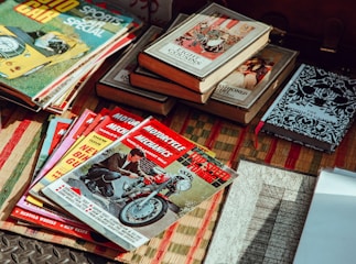 A collection of vintage magazines and books is spread out on a woven mat. The magazines feature topics like cars and motorcycles, with titles such as 'Motorcycle Mechanics' and 'Auto Car'. The books include older editions with classic designs and titles. The lighting gives a nostalgic feel to the scene.
