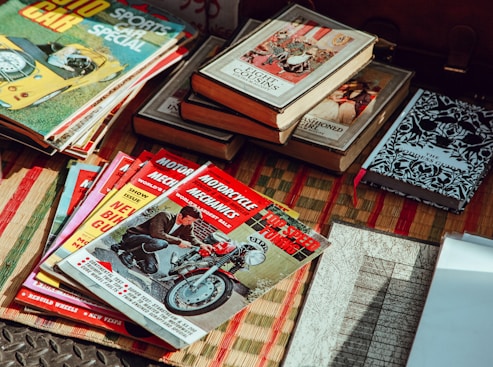 A collection of vintage magazines and books is spread out on a woven mat. The magazines feature topics like cars and motorcycles, with titles such as 'Motorcycle Mechanics' and 'Auto Car'. The books include older editions with classic designs and titles. The lighting gives a nostalgic feel to the scene.