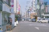 A busy urban street scene with cars parked along both sides and a few pedestrians walking on the sidewalks. The street is lined with various retail shops and businesses, featuring signage in Korean. The architecture consists of modern buildings with glass facades, and there are overhead utility lines and poles visible.