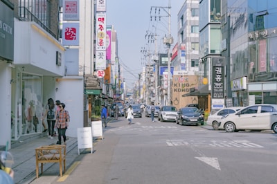 A busy urban street scene with cars parked along both sides and a few pedestrians walking on the sidewalks. The street is lined with various retail shops and businesses, featuring signage in Korean. The architecture consists of modern buildings with glass facades, and there are overhead utility lines and poles visible.