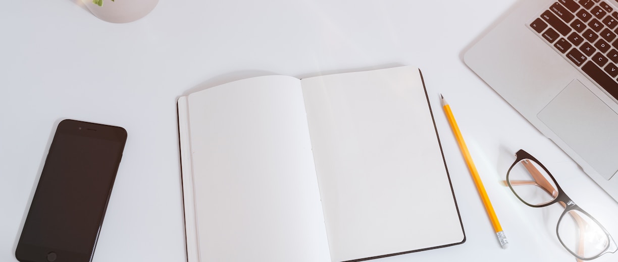 An open empty notebook on a white desk next to an iPhone and a MacBook