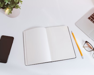 An open empty notebook on a white desk next to an iPhone and a MacBook