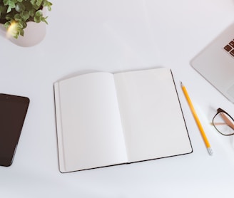 An open empty notebook on a white desk next to an iPhone and a MacBook