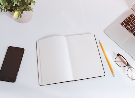 An open empty notebook on a white desk next to an iPhone and a MacBook