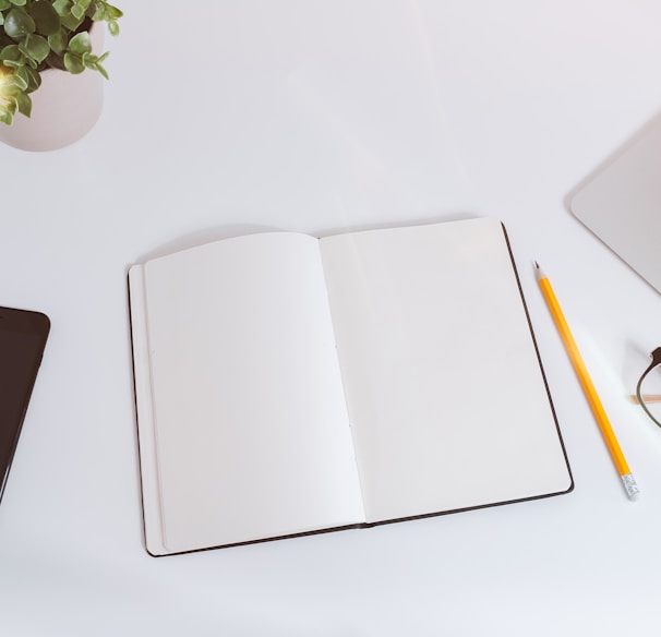 An open empty notebook on a white desk next to an iPhone and a MacBook