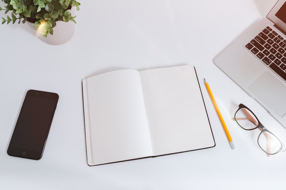 An open empty notebook on a white desk next to an iPhone and a MacBook