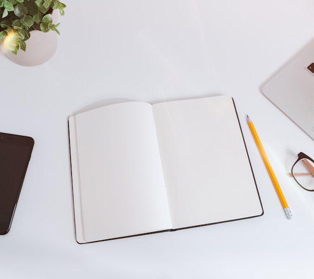 An open empty notebook on a white desk next to an iPhone and a MacBook