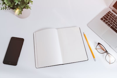 an open empty notebook on a white desk next to an iphone and a macbook book google meet background