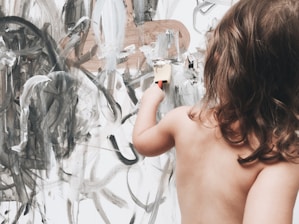 A young child with medium-length hair is painting an abstract design on a large canvas. The artwork consists of sweeping strokes of black, white, and grey, creating a chaotic yet artistic pattern. The child holds a paintbrush and seems focused on their creative expression.