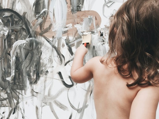 A young child with medium-length hair is painting an abstract design on a large canvas. The artwork consists of sweeping strokes of black, white, and grey, creating a chaotic yet artistic pattern. The child holds a paintbrush and seems focused on their creative expression.