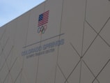 A building wall with the text 'COLORADO SPRINGS OLYMPIC TRAINING CENTER' and the Olympic rings is shown. An American flag is displayed above the text.