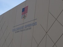A building wall with the text 'COLORADO SPRINGS OLYMPIC TRAINING CENTER' and the Olympic rings is shown. An American flag is displayed above the text.