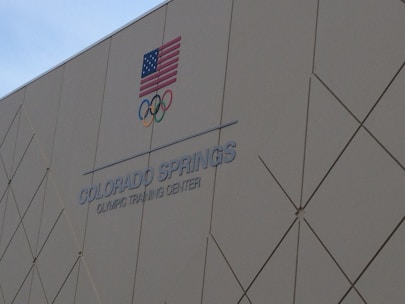 A building wall with the text 'COLORADO SPRINGS OLYMPIC TRAINING CENTER' and the Olympic rings is shown. An American flag is displayed above the text.