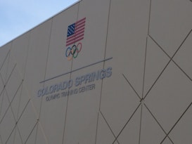 A building wall with the text 'COLORADO SPRINGS OLYMPIC TRAINING CENTER' and the Olympic rings is shown. An American flag is displayed above the text.