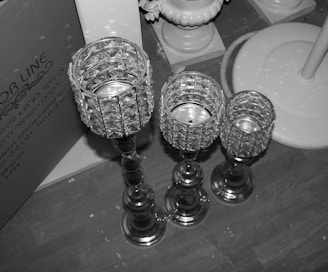 Three intricately designed candle holders of varying heights are displayed on a wooden floor. They feature crystal-like cups and metallic stems. A partially visible box with text in Hebrew and English rests in the background on the left side.