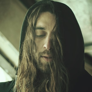 A person with long hair and a beard, wearing a dark hooded garment, has their eyes closed in a calming or introspective manner. The lighting is soft, creating a serene and contemplative atmosphere.