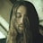 A person with long hair and a beard, wearing a dark hooded garment, has their eyes closed in a calming or introspective manner. The lighting is soft, creating a serene and contemplative atmosphere.