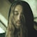 A person with long hair and a beard, wearing a dark hooded garment, has their eyes closed in a calming or introspective manner. The lighting is soft, creating a serene and contemplative atmosphere.