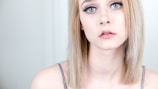 A young woman with light blonde hair and pale skin looks directly at the camera, with a neutral to slightly melancholic expression. She has green eyes and wears subtle makeup, including mascara and light pink lipstick. She is dressed in a thin-strapped top and is positioned against a plain, light-colored background.