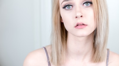 A young woman with light blonde hair and pale skin looks directly at the camera, with a neutral to slightly melancholic expression. She has green eyes and wears subtle makeup, including mascara and light pink lipstick. She is dressed in a thin-strapped top and is positioned against a plain, light-colored background.