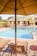 A poolside scene featuring lounge chairs under a large beige outdoor umbrella. The swimming pool is bordered by a brick patio and a building with multiple windows and outdoor seating. There is a potted palm tree next to the building, which adds a touch of greenery to the area.