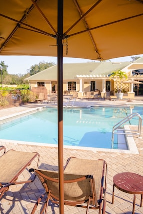 A poolside scene featuring lounge chairs under a large beige outdoor umbrella. The swimming pool is bordered by a brick patio and a building with multiple windows and outdoor seating. There is a potted palm tree next to the building, which adds a touch of greenery to the area.