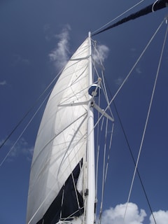 A large white sail with a sturdy mast set against a clear blue sky. The sail is hoisted high, capturing the wind on a calm day. The rigging and lines are visible, supporting the structure of the sail.