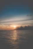 A serene ocean scene with gentle waves and the sun setting behind distant mountains. The sky is adorned with wispy clouds and a jet stream, creating a peaceful and picturesque atmosphere.