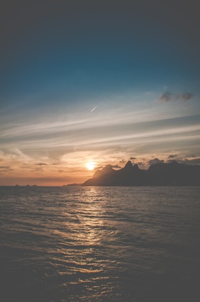 A serene ocean scene with gentle waves and the sun setting behind distant mountains. The sky is adorned with wispy clouds and a jet stream, creating a peaceful and picturesque atmosphere.
