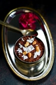 A decorative silver plate features a traditional Indian dessert garnished with cashew nuts and silver leaf. A small spoon and a vibrant red flower accompany the arrangement, enhancing the elegant presentation.