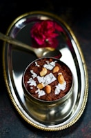 A decorative silver plate features a traditional Indian dessert garnished with cashew nuts and silver leaf. A small spoon and a vibrant red flower accompany the arrangement, enhancing the elegant presentation.