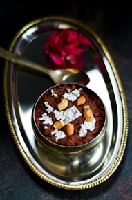 A decorative silver plate features a traditional Indian dessert garnished with cashew nuts and silver leaf. A small spoon and a vibrant red flower accompany the arrangement, enhancing the elegant presentation.