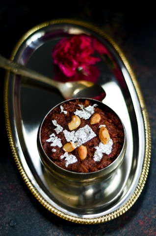 A decorative silver plate features a traditional Indian dessert garnished with cashew nuts and silver leaf. A small spoon and a vibrant red flower accompany the arrangement, enhancing the elegant presentation.