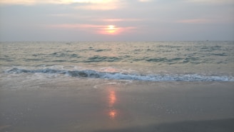 A serene ocean scene with gentle waves lapping against the sandy shore. The sky is tinged with soft hues as the sun sets on the horizon, casting a warm reflection over the water.