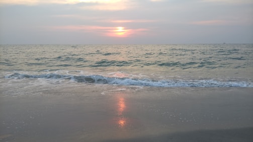 A serene ocean scene with gentle waves lapping against the sandy shore. The sky is tinged with soft hues as the sun sets on the horizon, casting a warm reflection over the water.