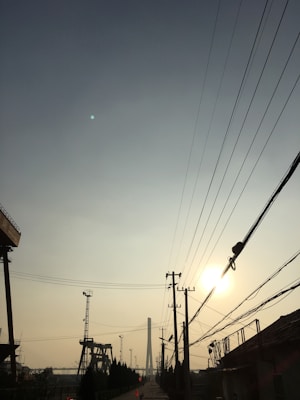 Telecommunication poles and wires stretch across the sky, converging towards the horizon where a tall tower stands. The scene captures a silhouette of industrial structures with the sun setting in the distance, casting a warm glow over the landscape.