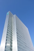 A tall, modern skyscraper with a grid-like facade of windows and a metallic exterior. The building rises against a clear blue sky, with a logo displayed near its rooftop.