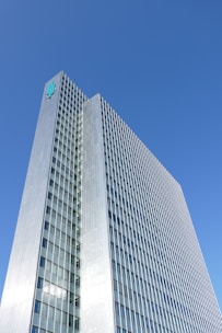 A tall, modern skyscraper with a grid-like facade of windows and a metallic exterior. The building rises against a clear blue sky, with a logo displayed near its rooftop.
