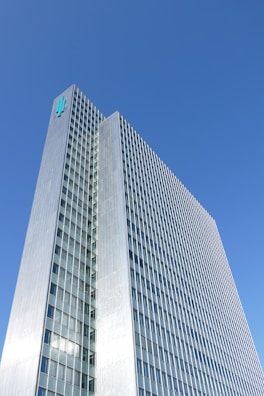 A tall, modern skyscraper with a grid-like facade of windows and a metallic exterior. The building rises against a clear blue sky, with a logo displayed near its rooftop.